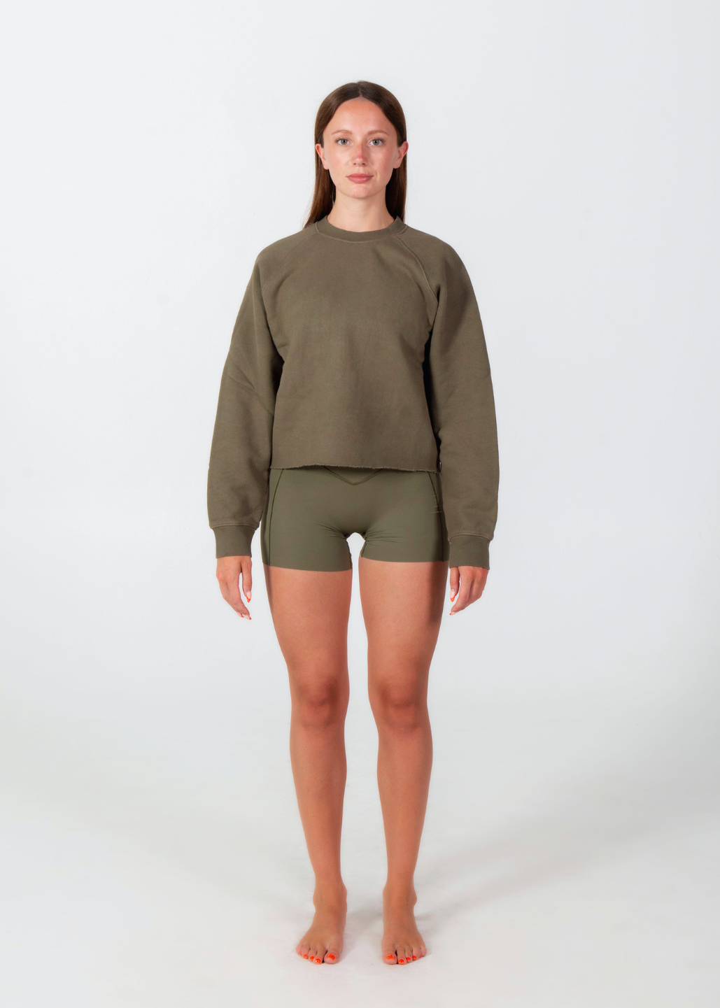 A woman stands facing forward in a studio wearing an olive green long-sleeve cropped sweatshirt and matching olive green shorts against a plain light gray background.