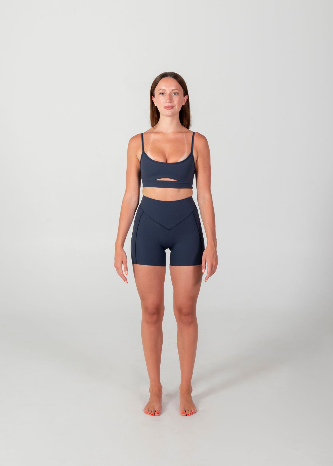 A woman stands barefoot against a light gray studio background wearing a matching dark navy activewear set consisting of a sports bra with a horizontal cutout and high-waisted shorts with side pockets.