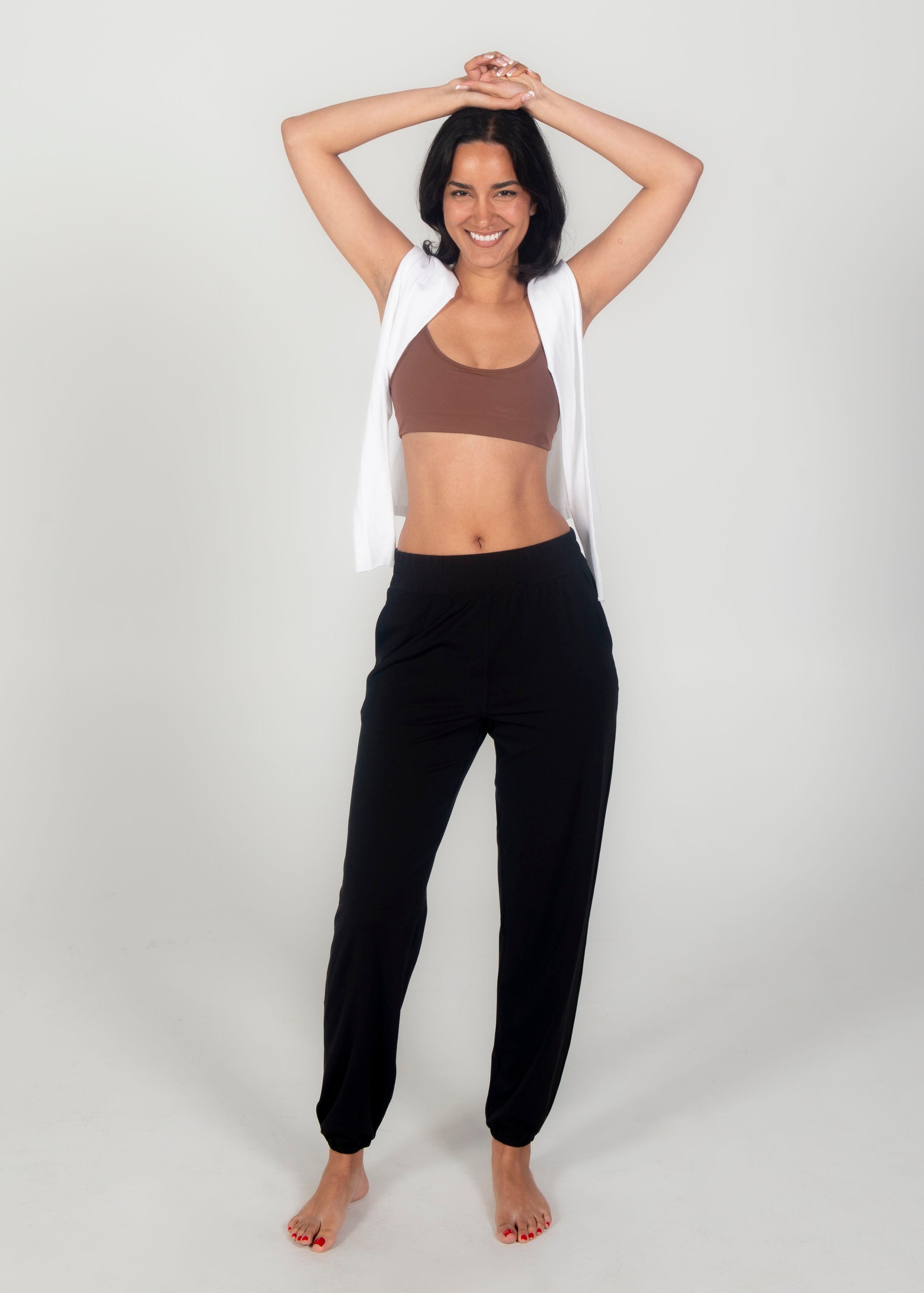 Woman in brown sports bra and black leggings with a white towel over her shoulder on a light gray background