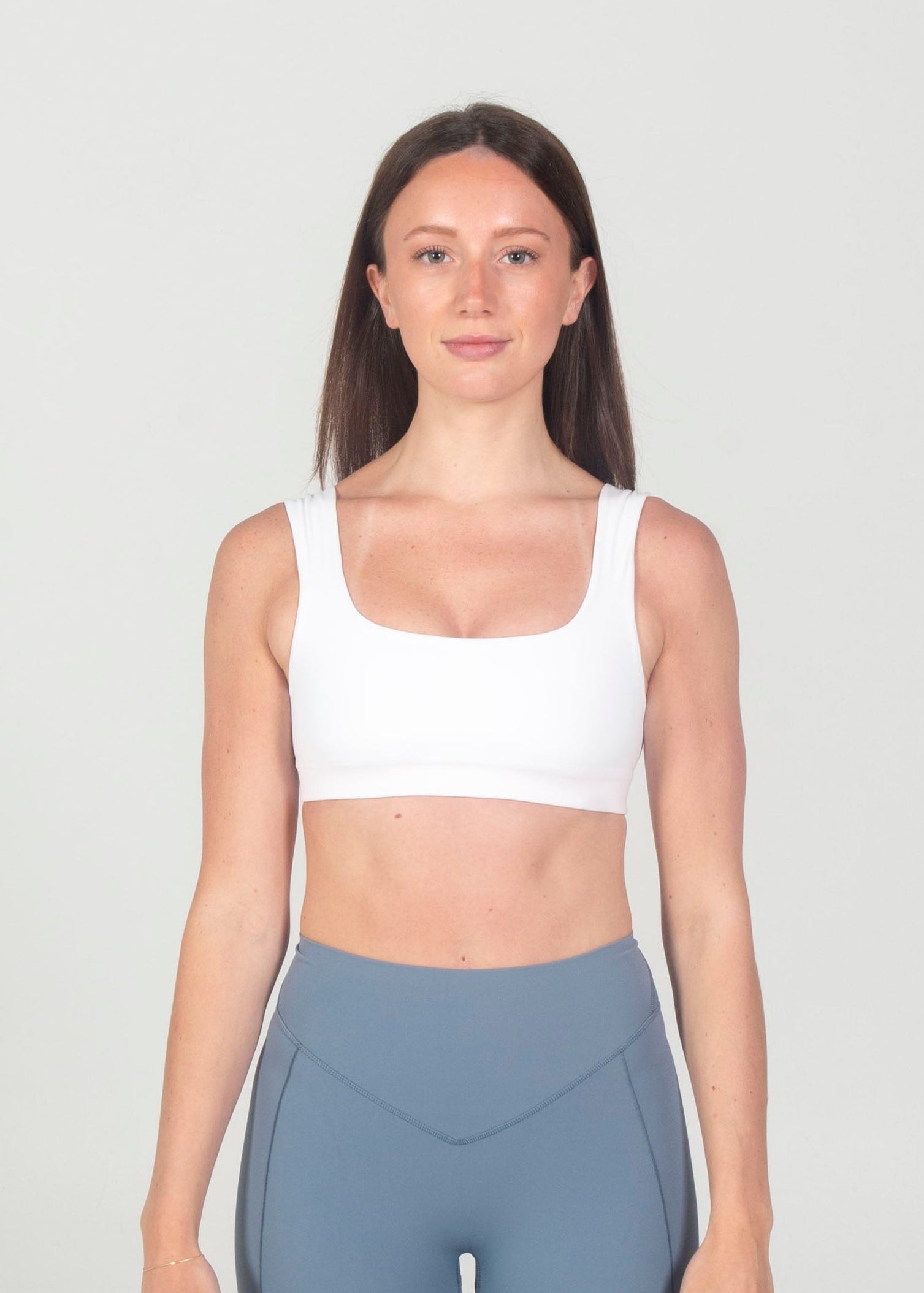 Woman wearing a white sports bra and gray leggings on a plain background